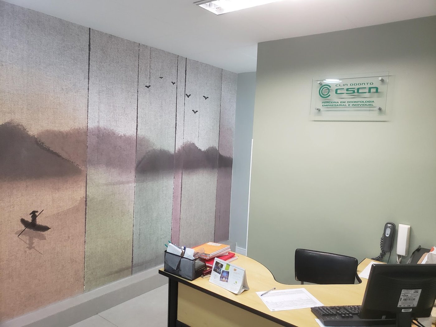 Recepção da clínica, com balcão de atendimento, painel de parede com paisagem e placa da CSCN